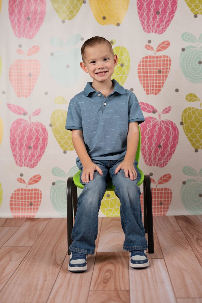 Smiling preschool boy posing for a school portrait in front of a colorful apple backdrop.